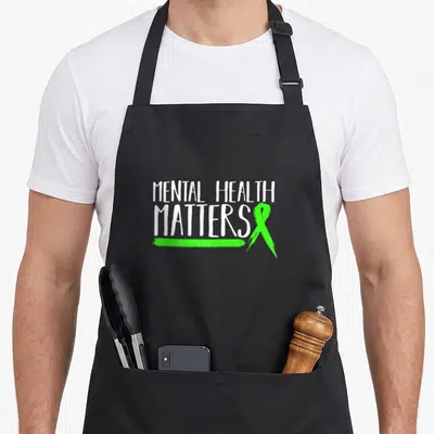 Cute matter mental health awareness apron - White stacked text MENTAL HEALTH MATTERS with green ribbon and brush stroke — shown on a black cotton canvas apron against a white background
