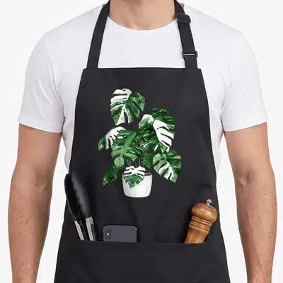 Variegated plant apron - Watercolor monstera with green and white variegated leaves in a white pot — shown on a black cotton canvas apron against a white background