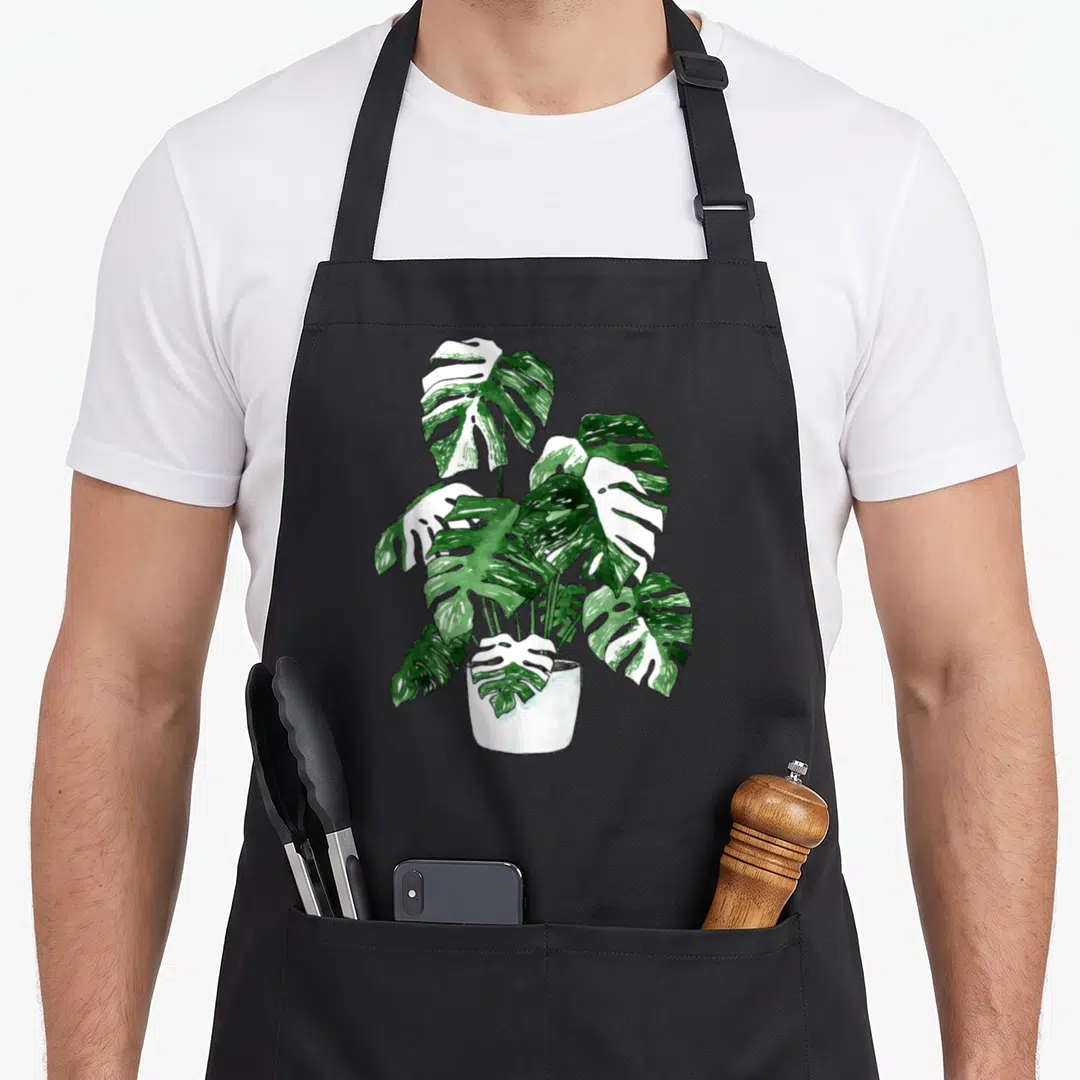 Variegated plant apron - Watercolor monstera with green and white variegated leaves in a white pot — shown on a black cotton canvas apron against a white background