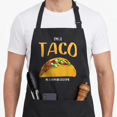 Cosplay halloween apron - Cartoon taco illustration with text I'M A TACO IN A HUMAN COSTUME — shown on a black cotton canvas apron against a white background