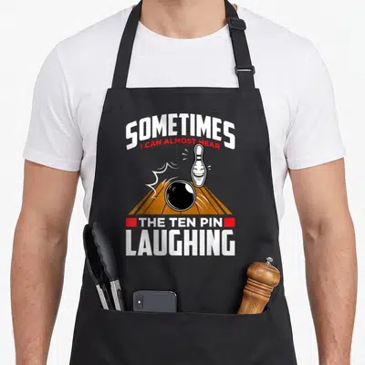 Hear pins for aprons - Laughing bowling pin and ball on orange lane with sarcastic text — shown on a black cotton canvas apron against a white background