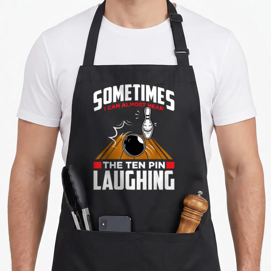 Hear pins for aprons - Laughing bowling pin and ball on orange lane with sarcastic text — shown on a black cotton canvas apron against a white background
