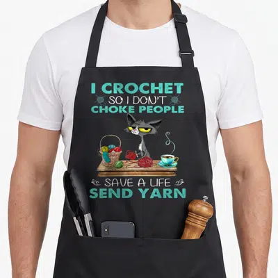 Choke crochet apron - Cartoon grey cat at table with yarn basket and tea under teal slogan text — shown on a black cotton canvas apron against a white background