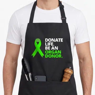 Donate organ apron - Green awareness ribbon next to white and green Donate Life Be an Organ Donor text — shown on a black cotton canvas apron against a white background