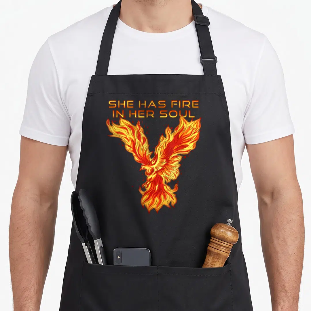 She bird apron - Cartoon phoenix in orange yellow and red below stacked text SHE HAS FIRE IN HER SOUL with red sparks — shown on a black cotton canvas apron against a white background