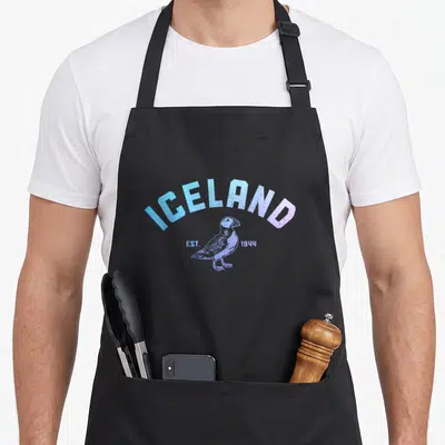 Iceland puffin icelandic apron - Distressed blue and purple ICELAND EST 1944 text arching over a standing puffin — shown on a black cotton canvas apron against a white background