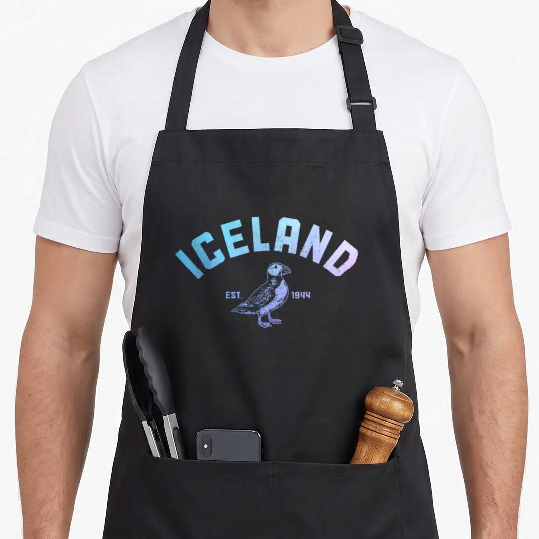 Iceland puffin icelandic apron - Distressed blue and purple ICELAND EST 1944 text arching over a standing puffin — shown on a black cotton canvas apron against a white background