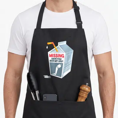 Amputee milk aprons - Cartoon milk carton with red text MISSING HAVE YOU SEEN MY LEG and prosthetic leg graphic — shown on a black cotton canvas apron against a white background