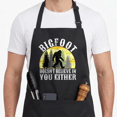 Either bigfoot apron - Bigfoot silhouette in yellow sun with three pine trees and arched white text — shown on a black cotton canvas apron against a white background