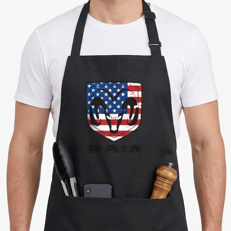 Dodge ram apron - Black ram head on red white blue shield with RAM text below — shown on a black cotton canvas apron against a white background