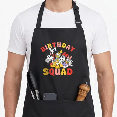 Birthday friends cooking apron - Cartoon Mickey Minnie Donald Goofy Pluto cluster under arched BIRTHDAY SQUAD text — shown on a black cotton canvas apron against a white background