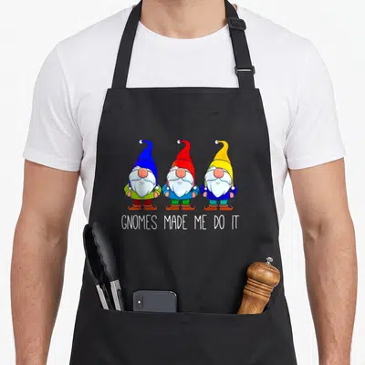 Garden funny gnome apron - Three cartoon gnomes with blue, red, and yellow hats standing above white text — shown on a black cotton canvas apron against a white background