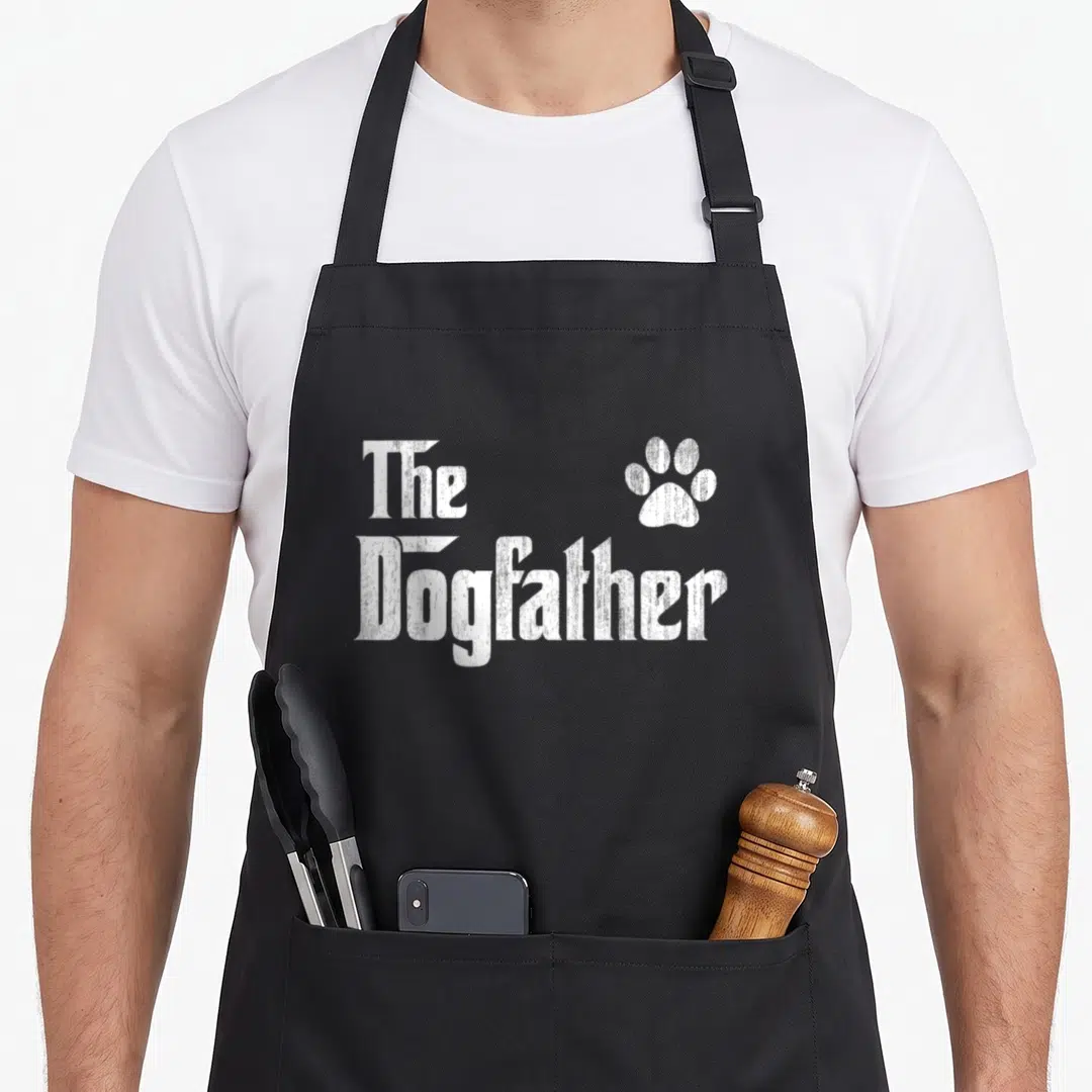 Cute dogfather dog dad apron - White distressed text reading The Dogfather beside a paw print — shown on a black cotton canvas apron against a white background