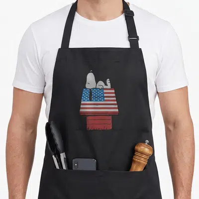 Funny peanut lover apron - Snoopy lying on red doghouse with USA flag roof pattern in cartoon style — shown on a black cotton canvas apron against a white background