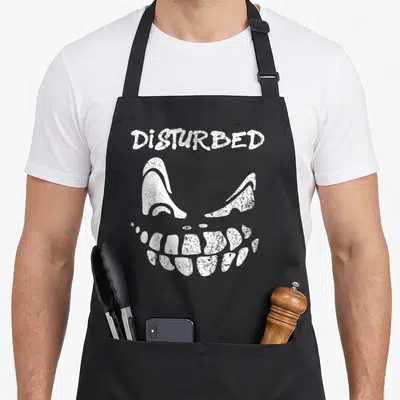 Disturbed face apron - White distressed text DISTURBED above stylized eyes and grinning skull mouth — shown on a black cotton canvas apron against a white background