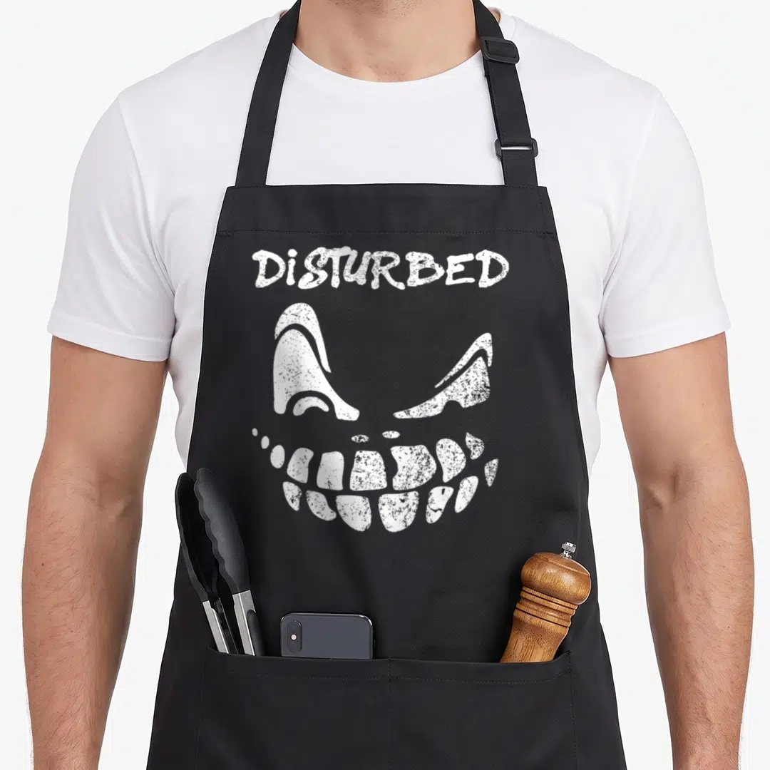 Disturbed face apron - White distressed text DISTURBED above stylized eyes and grinning skull mouth — shown on a black cotton canvas apron against a white background
