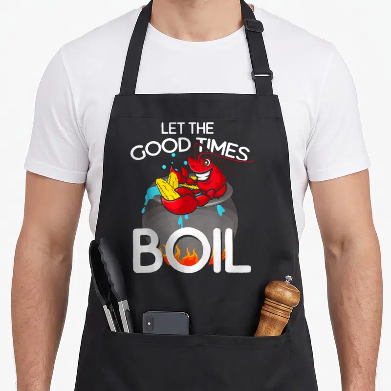 Crawdaddy crawfish boil apron - Cartoon red crawfish in grey pot with corn and text Let The Good Times Boil — shown on a black cotton canvas apron against a white background
