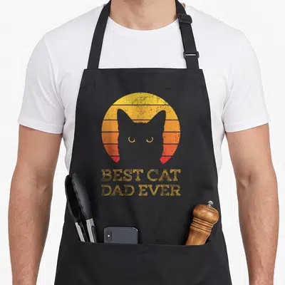 Daddy cat dad apron - Grey cat silhouette in front of yellow orange red striped sun with BEST CAT DAD EVER text — shown on a black cotton canvas apron against a white background