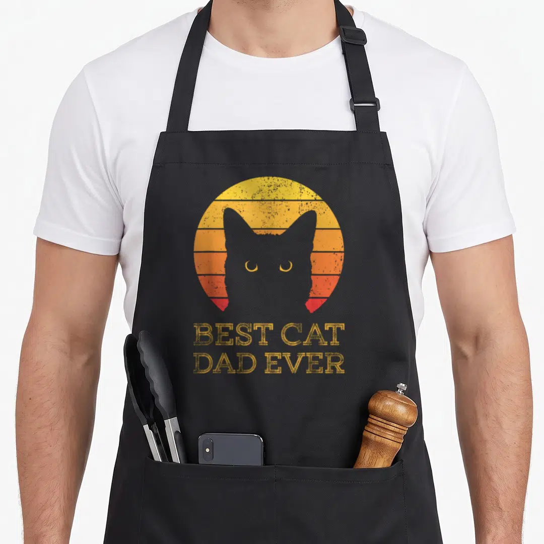 Daddy cat dad apron - Grey cat silhouette in front of yellow orange red striped sun with BEST CAT DAD EVER text — shown on a black cotton canvas apron against a white background