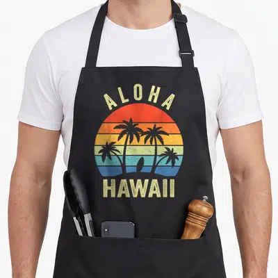 Aloha hawaiian apron - Retro sunset with palm trees, surfboard, and ALOHA HAWAII text in stripes — shown on a black cotton canvas apron against a white background