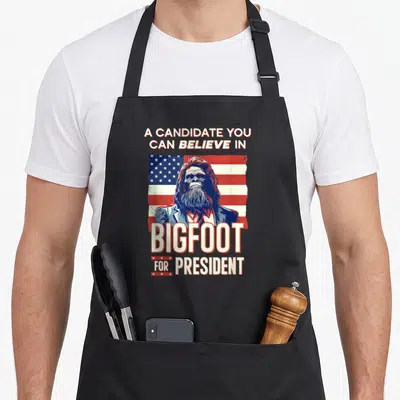 President bigfoot apron - Bigfoot in a suit with red white blue campaign text and distressed flag background — shown on a black cotton canvas apron against a white background