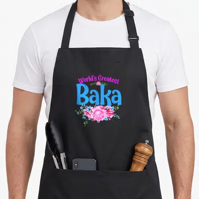 Greatest croatian aprons - Arched pink text World's Greatest above blue Baka with pink and blue flower cluster — shown on a black cotton canvas apron against a white background