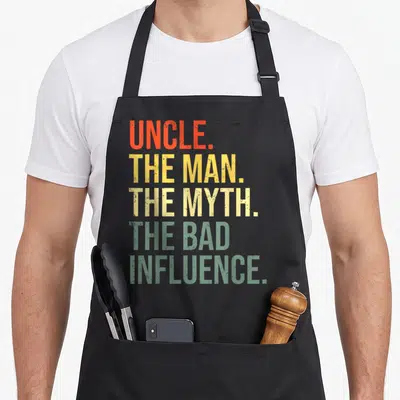 Uncle funcle apron - Stacked distressed text reading UNCLE THE MAN THE MYTH THE BAD INFLUENCE in orange yellow and green — shown on a black cotton canvas apron against a white background