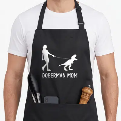 Dobe dog apron - White silhouette of woman walking dinosaur with DOBERMAN MOM text — shown on a black cotton canvas apron against a white background