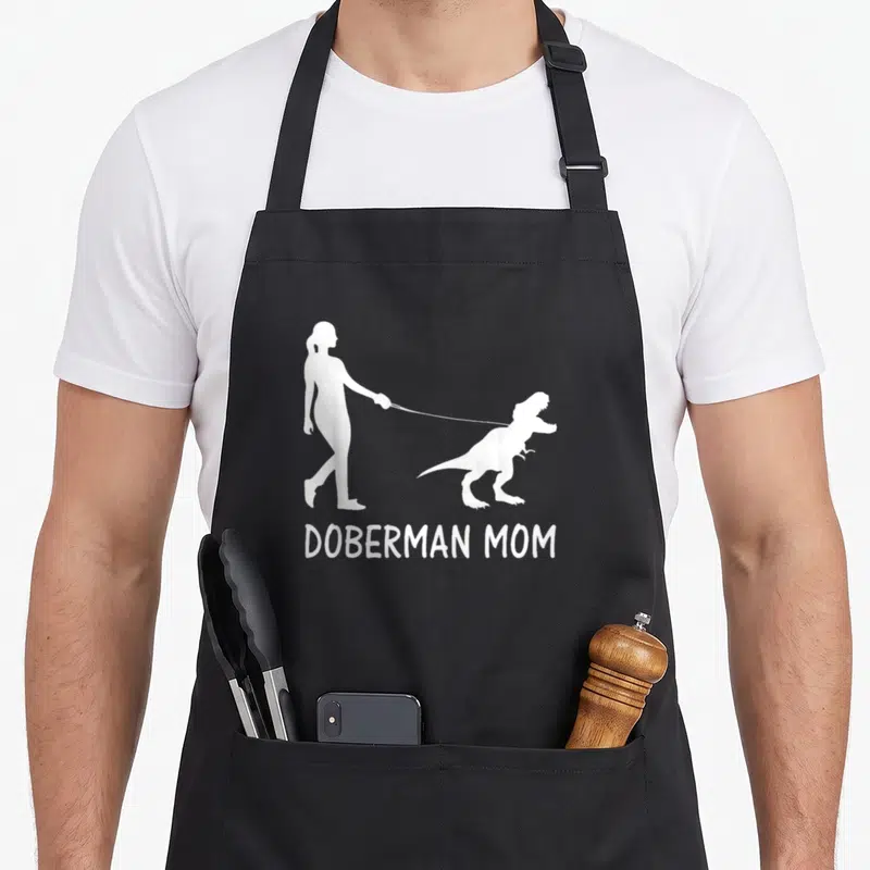 Dobe dog apron - White silhouette of woman walking dinosaur with DOBERMAN MOM text — shown on a black cotton canvas apron against a white background