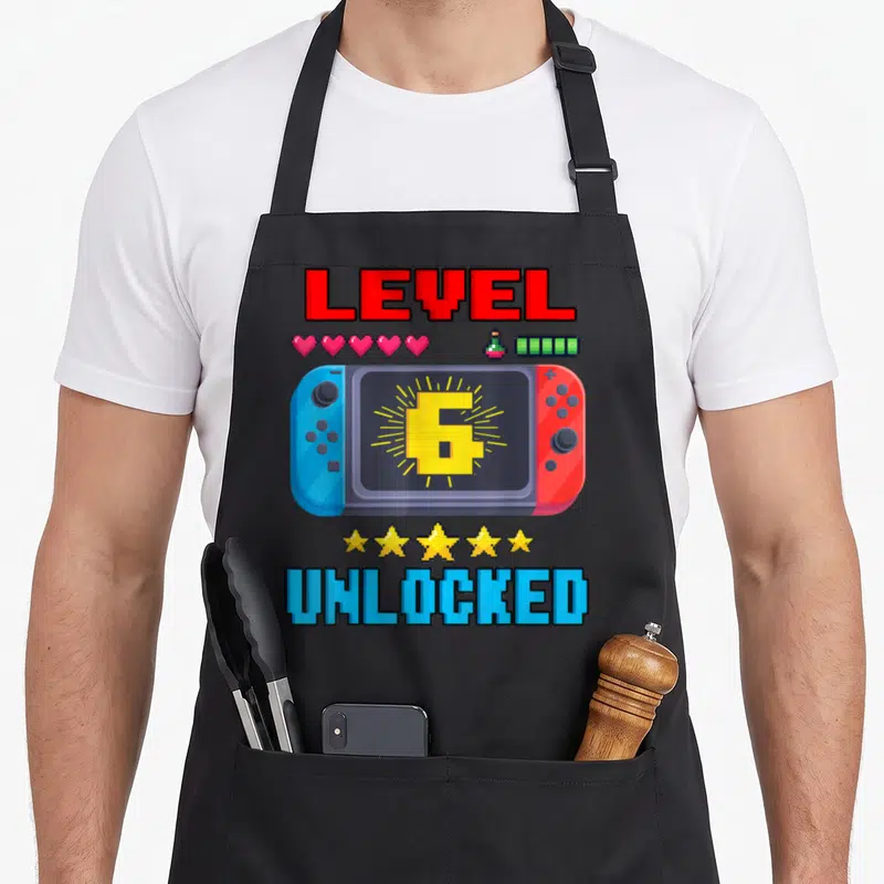 6th level birthday apron - Pixel handheld console with number 6 screen, pink hearts, yellow stars, red blue text — shown on a black cotton canvas apron against a white background