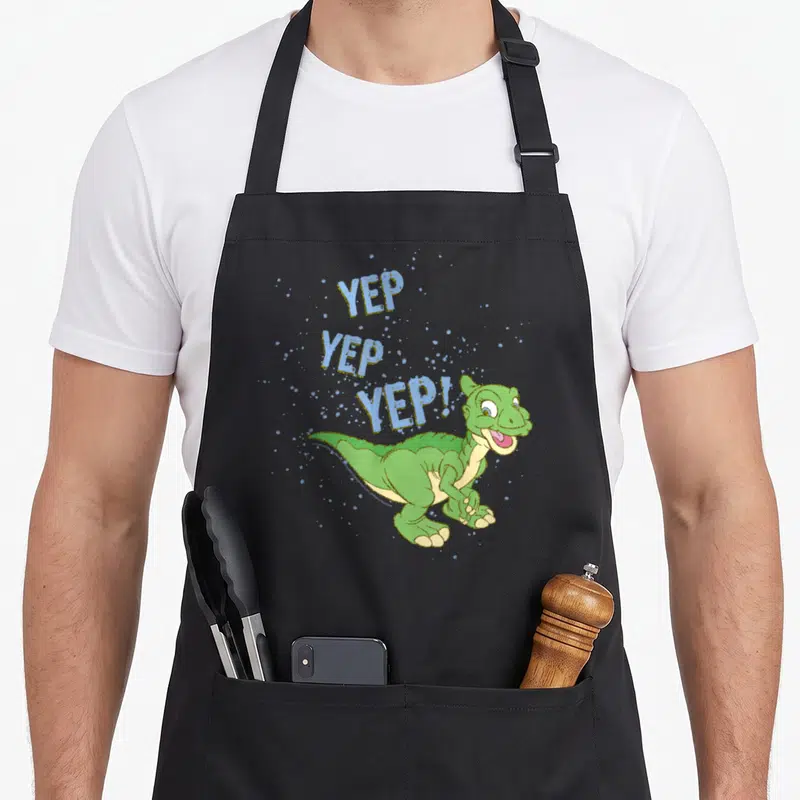 Before sprinkle apron - Green cartoon dinosaur with three stacked YEP words in light blue and gold splatters — shown on a black cotton canvas apron against a white background