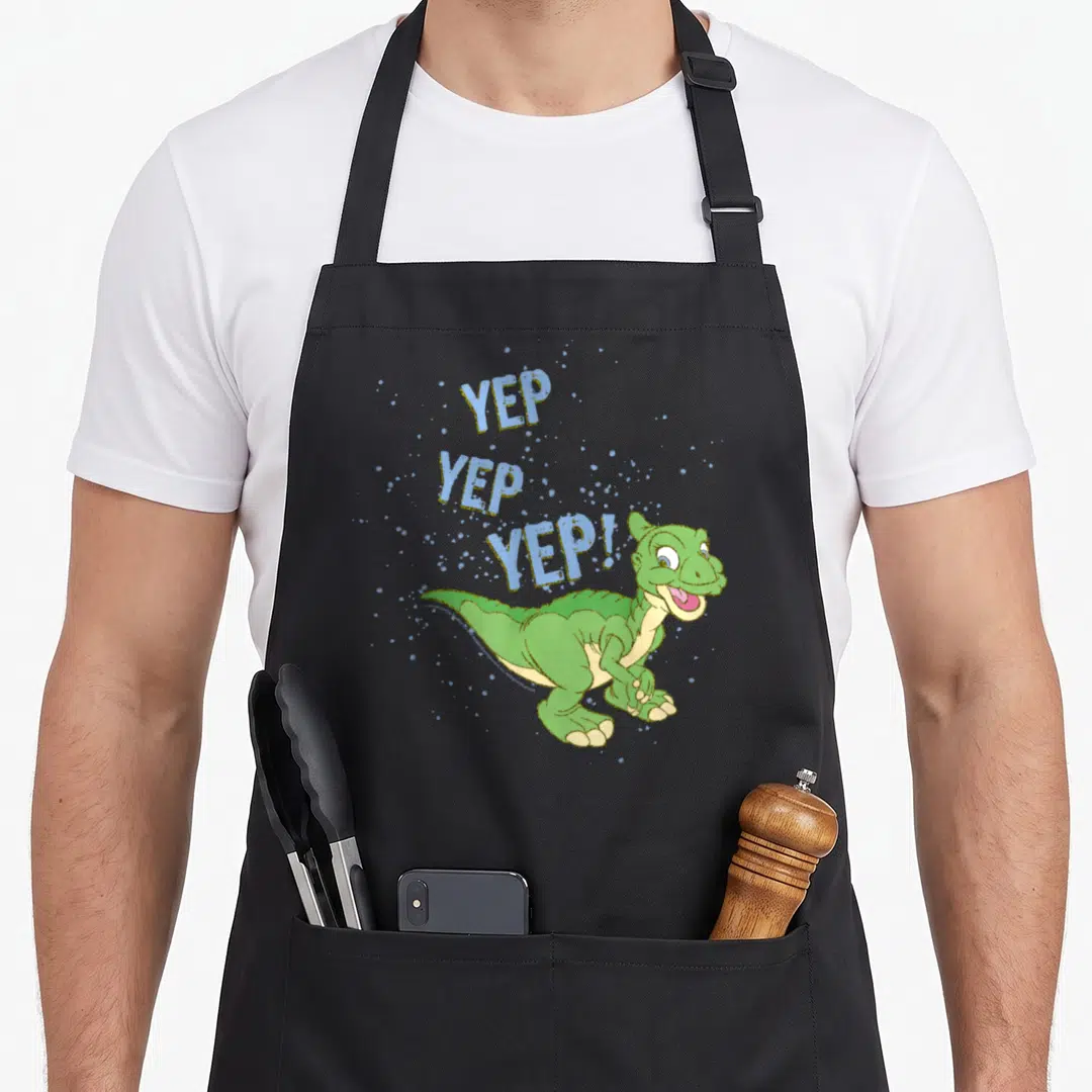 Before sprinkle apron - Green cartoon dinosaur with three stacked YEP words in light blue and gold splatters — shown on a black cotton canvas apron against a white background