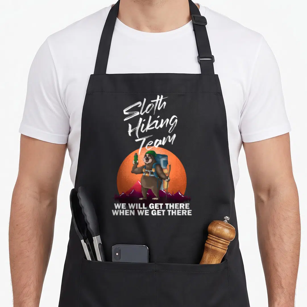 Backpacking sloth apron - Cartoon brown sloth with blue backpack and green hat in front of orange sun and purple — shown on a black cotton canvas apron against a white background