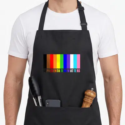 Tran pride apron - vertical stripes in black brown red orange yellow green blue purple white light blue pink above — shown on a black cotton canvas apron against a white background