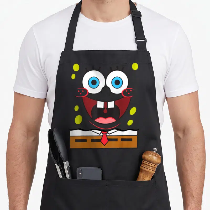 Smile general apron - SpongeBob face with wide mouth, red tie, brown pants, and green bubbles — shown on a black cotton canvas apron against a white background