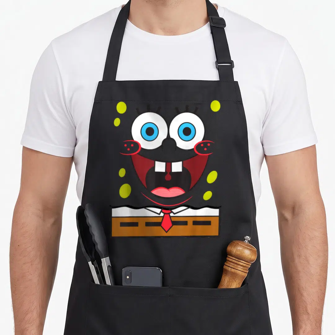 Smile general apron - SpongeBob face with wide mouth, red tie, brown pants, and green bubbles — shown on a black cotton canvas apron against a white background