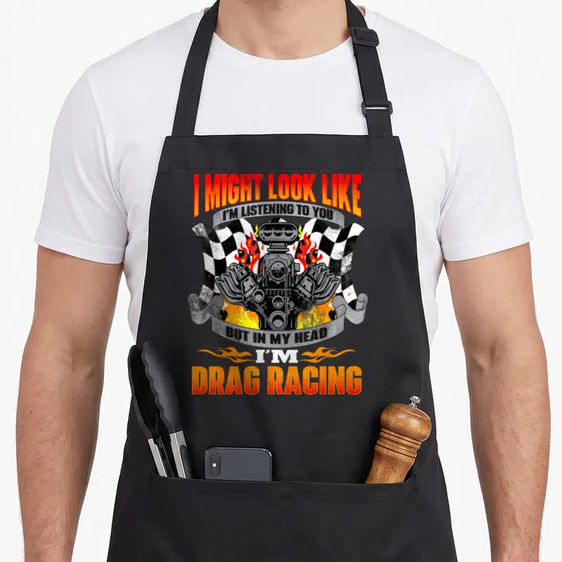 Racer car apron - Cartoon engine flanked by two checkered flags and fire with stacked text ribbons — shown on a black cotton canvas apron against a white background