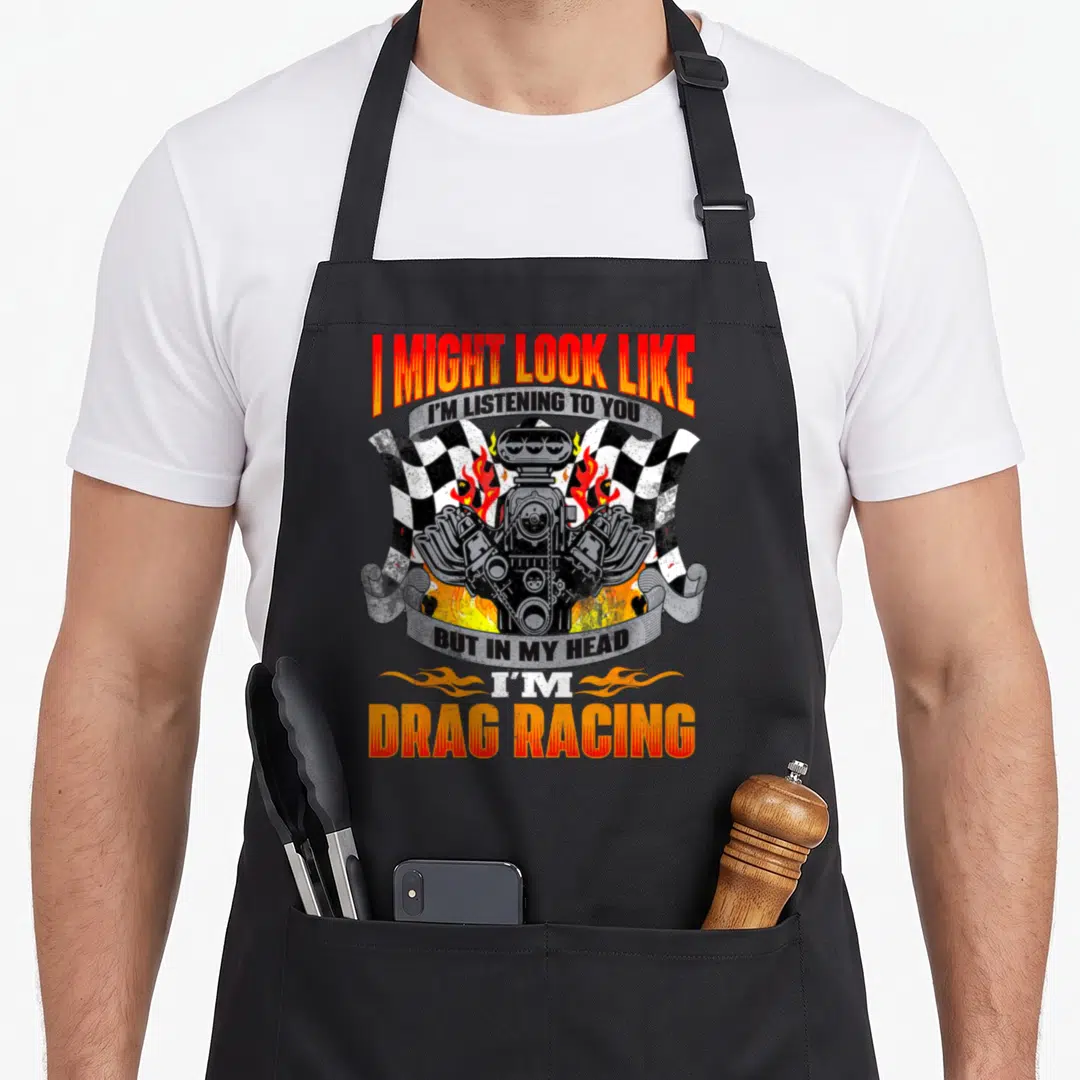 Racer car apron - Cartoon engine flanked by two checkered flags and fire with stacked text ribbons — shown on a black cotton canvas apron against a white background