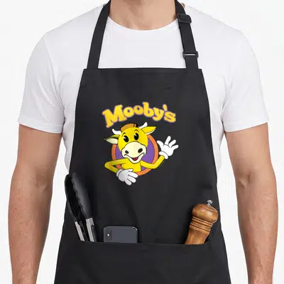 Bob jay lover apron - Yellow cartoon cow waving inside orange-purple circle under arched Mooby's text — shown on a black cotton canvas apron against a white background