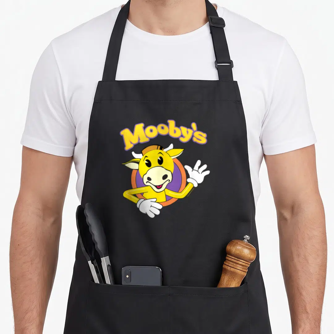 Bob jay lover apron - Yellow cartoon cow waving inside orange-purple circle under arched Mooby's text — shown on a black cotton canvas apron against a white background
