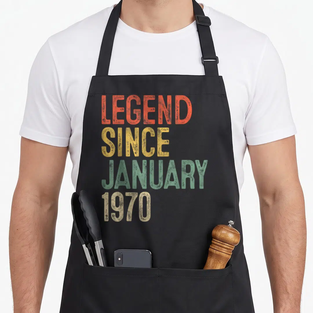 January 52nd birthday apron - Stacked distressed text reading LEGEND SINCE JANUARY 1970 in red yellow green and beige — shown on a black cotton canvas apron against a white background