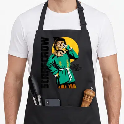 Funny scarecrow wizard of oz apron - Green cartoon scarecrow in front of yellow sun with vertical SCARECROW text — shown on a black cotton canvas apron against a white background