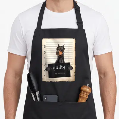 Shot dog apron - Doberman holding a black sign reading guilty against a beige height chart — shown on a black cotton canvas apron against a white background