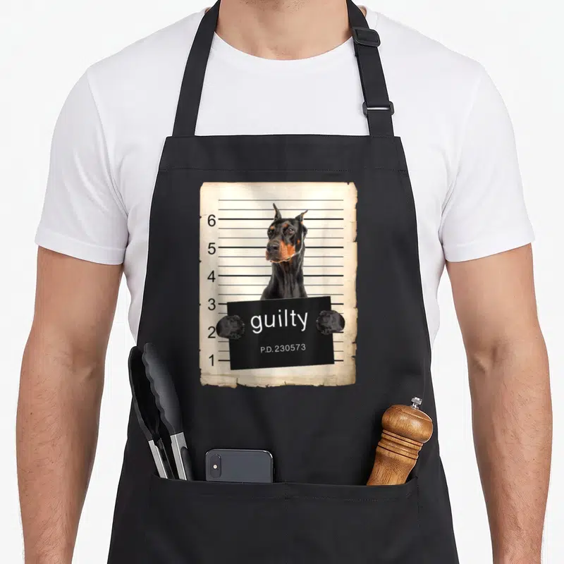 Shot dog apron - Doberman holding a black sign reading guilty against a beige height chart — shown on a black cotton canvas apron against a white background