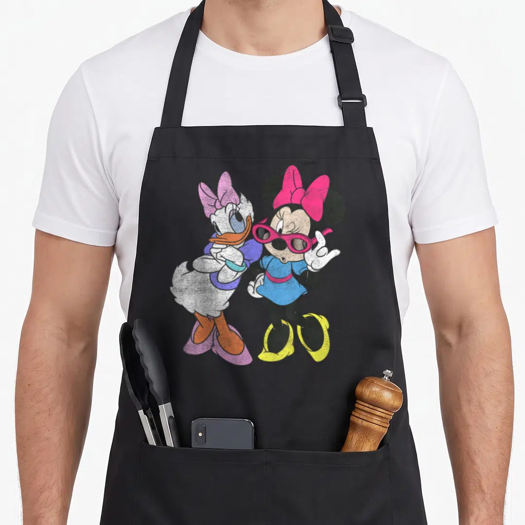 Daisy friends apron - Minnie Mouse in sunglasses and Daisy Duck standing together in pink, blue, and yellow cartoon art — shown on a black cotton canvas apron against a white background
