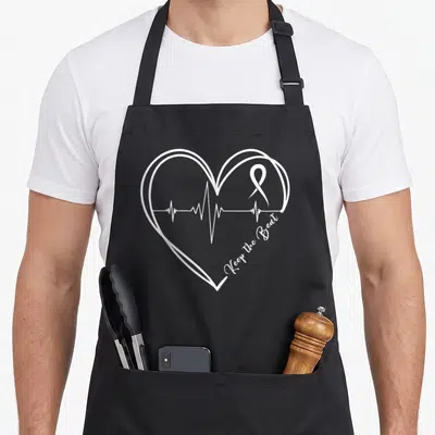 Heartbeat disease awareness apron - White line art heart outline with EKG line and Keep the Beat script text — shown on a black cotton canvas apron against a white background