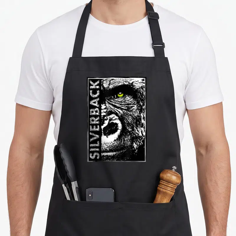 Silverback face apron - Distressed black white gorilla face with vertical SILVERBACK text and yellow eye — shown on a black cotton canvas apron against a white background