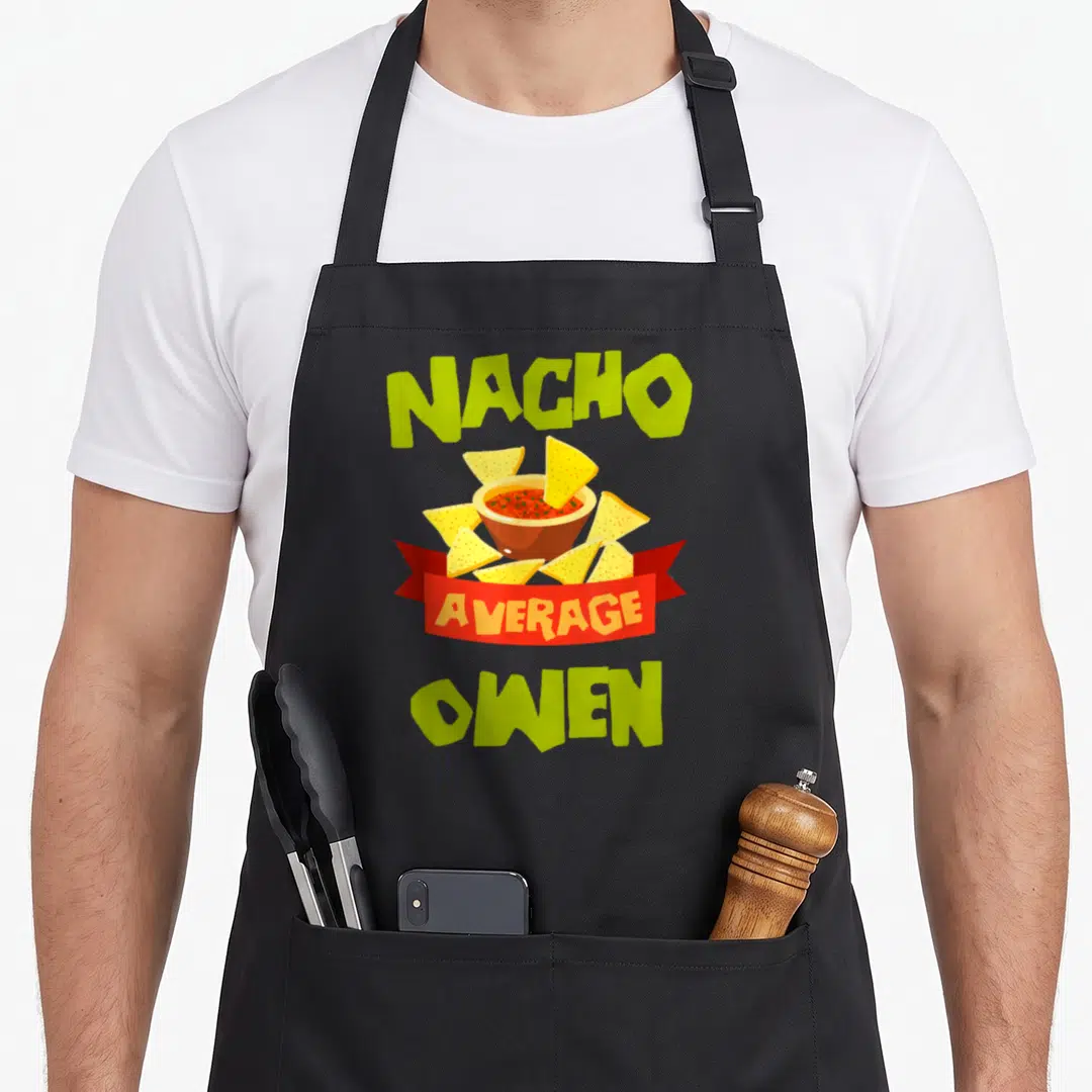 Mexican father's day aprons - Nacho Average Owen cartoon text with bowl of red dip and yellow chips on red ribbon — shown on a black cotton canvas apron against a white background