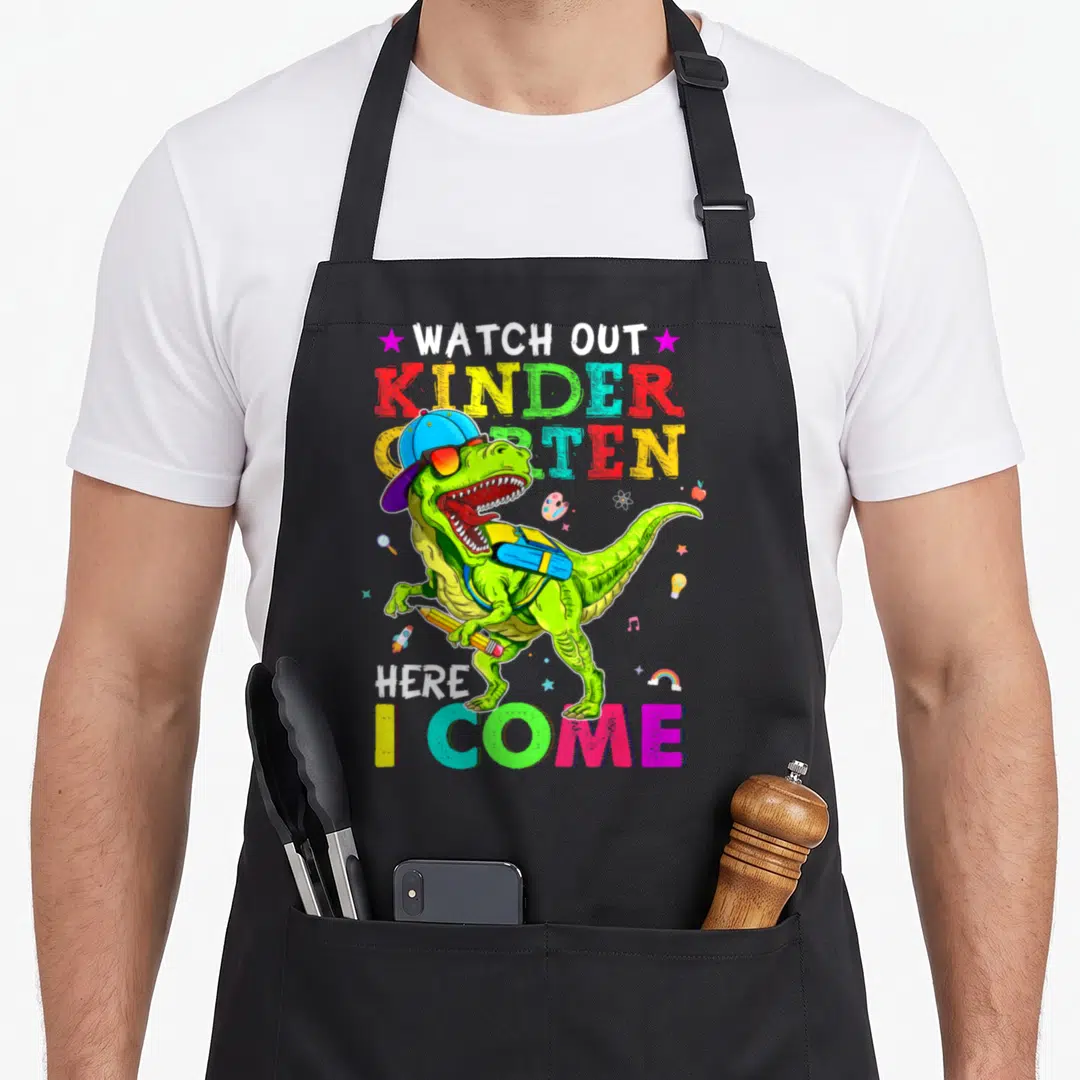 Watch dinosaur apron - Cartoon T-Rex wearing hat and glasses holding pencil with school icons — shown on a black cotton canvas apron against a white background