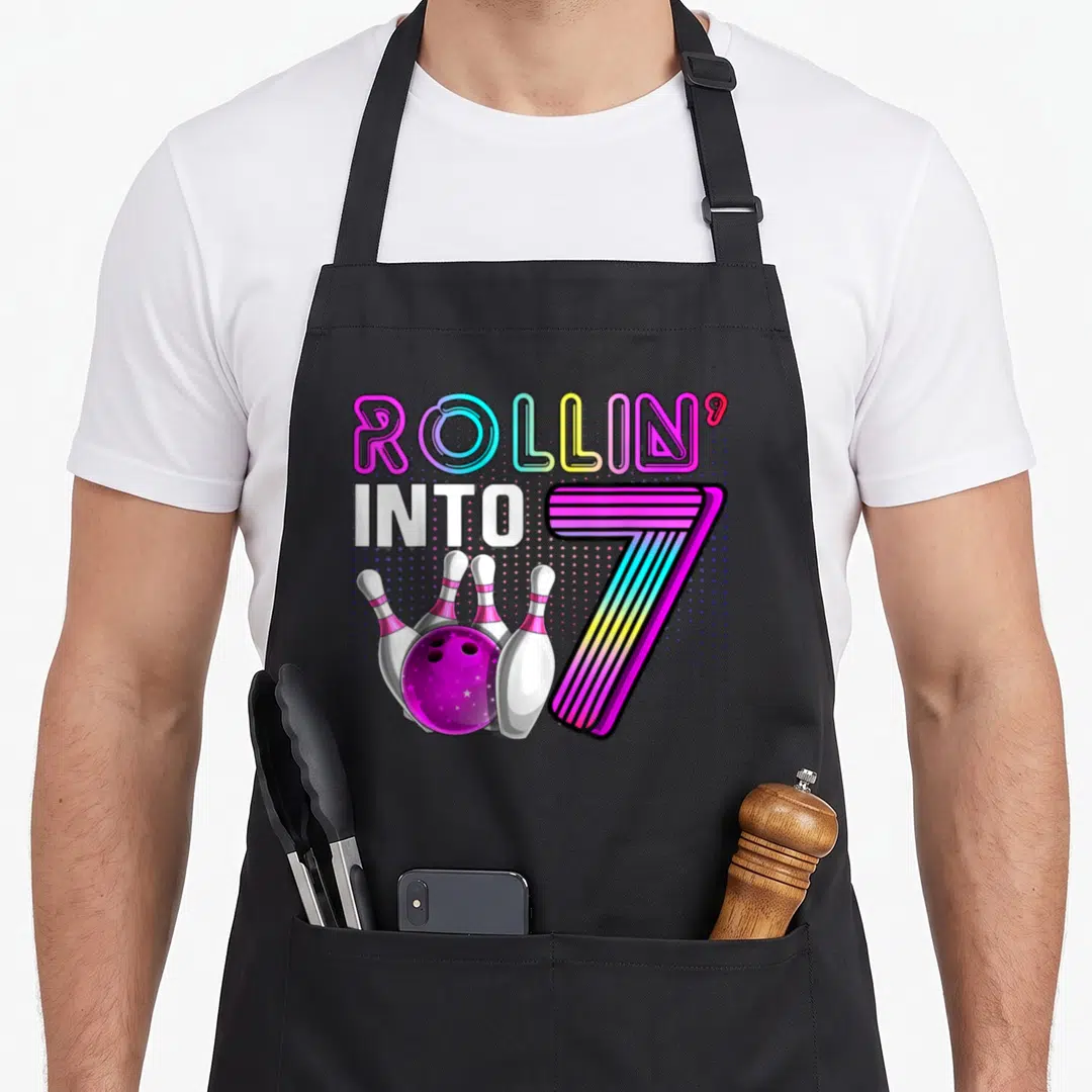 Bowling 7th birthday apron - Rollin into 7 text with cartoon bowling ball and pins in purple pink and blue — shown on a black cotton canvas apron against a white background